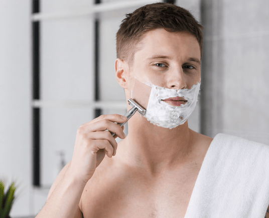 Man shaving his face with a razor and shaving cream in a bathroom setting.