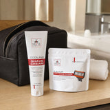 Shave Essentials grooming kit on a wooden bathroom counter, featuring a black zippered travel case, a white tube of Cooling Shave Cream, a white cartridge razor pouch, and a rolled white towel.